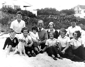 The Kennedy Family at Hyannis Port, 1931. L-R: Robert Kennedy, John F. Kennedy, Eunice Kennedy, Jean Kennedy (on lap of) Joseph P. Kennedy Sr., Rose Fitzgerald Kennedy (behind) Patricia Kennedy, Kathleen Kennedy, Joseph P. Kennedy Jr. (behind) Rosemary Kennedy. Dog in foreground is "Buddy". Photograph by Richard Sears in the John F. Kennedy Presidential Library and Museum, Boston.