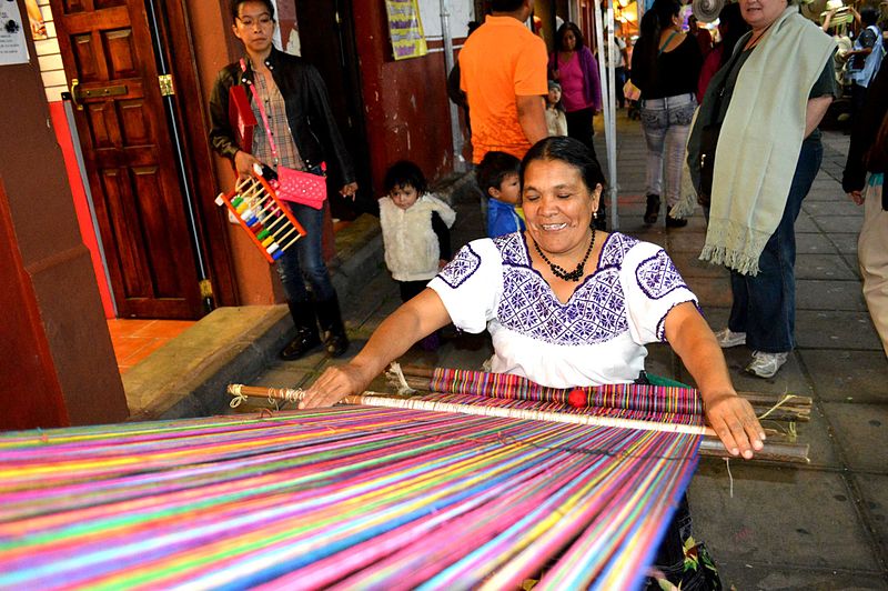 Indigenous women weaving on a backstrap loom during the Tianguis de Domingo de Ramos in Uruapan, Michoacan, Mexico. Attribution: AlejandroLinaresGarcia